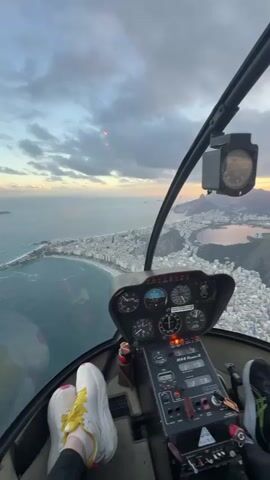 The video displays an aerial view from the interior of a helicopter cockpit, looking out over a coastal city at what appears to be sunset or sunrise. In the immediate foreground, the helicopter's instrument panel is visible, featuring various gauges, switches, and a digital display. Below the instrument panel, two pairs of feet are present, one wearing white sneakers with yellow laces and the other wearing black sneakers with white soles. Through the cockpit windows, a sprawling cityscape extends along a curved coastline with a sandy beach adjacent to a large body of water. Numerous buildings with light-colored roofs are densely packed across the urban area. In the distance, several large, green, tree-covered mountains are visible, with one prominent peak and a lake nestled behind it. The sky is partly cloudy, with warm orange and yellow hues on the horizon contrasting with the grey clouds above.