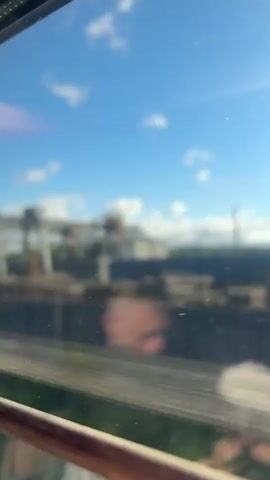 A person is seen through a train window with a can of drink on the table in front of them. Outside the train, there's an industrial area with large cranes and stacks of materials. The sky is blue with scattered clouds. The train is moving. The image is somewhat blurry due to the train window.