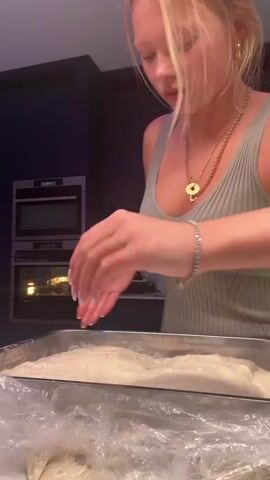 A young woman with blonde hair and fair skin is preparing dough in a kitchen. She is wearing a gray tank top, gold jewelry, and a silver bracelet. Her nails are painted white. She is working with a large mass of dough in a metal baking pan lined with plastic wrap. She repeatedly presses, folds, and manipulates the dough with her hands. The kitchen has dark cabinets, and two ovens are visible in the background. The lower oven appears to be in use.
