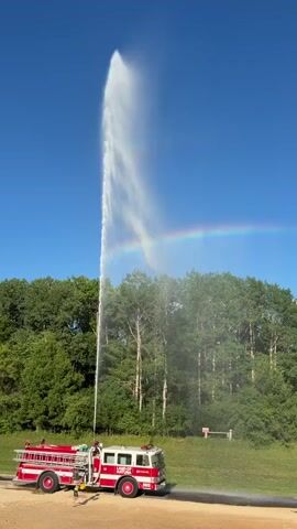 A red and white fire truck sprays a powerful jet of water high into the air against a bright blue sky. A faint rainbow is visible in the mist created by the water. A line of trees forms a backdrop behind the fire truck, and a few people, appearing to be children, are visible in the foreground on a sandy area.