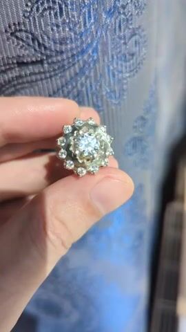 A person's hand holds a ring with a large center stone and smaller stones around it. The background is a blue textured fabric.