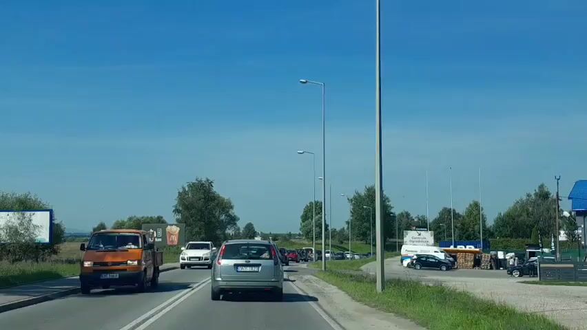 A clear, sunny day with a bright blue sky is depicted as the camera travels forward on a road. The road is a two-lane highway with a white line dividing the lanes. Several vehicles are visible on the road. The closest vehicle in the foreground is an orange van, followed by a silver hatchback. Behind the silver hatchback are several other cars, forming a moderate traffic flow. On the left side of the road, there is a grassy shoulder with trees and bushes. A white SUV is in the left lane, passing the orange van. A billboard with an advertisement for food is also visible on the left. On the right side of the road, there is a grassy shoulder with streetlights and a building with parking. Several cars are parked in the lot, and some structures that appear to be shops or stalls are set up. The landscape is generally rural, with trees and fields visible in the distance. The video appears to be filmed from the perspective of a passenger in a car.