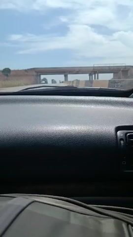 The view from inside a moving vehicle, most likely a car, as it approaches a concrete overpass bridge on a road or highway. The interior of the car is visible, focusing on the dashboard, which is a dark color with a textured surface. Below the dashboard, a part of a seatbelt is visible. Through the windshield, the road ahead is seen, leading towards the overpass. Several large trucks and at least one pedestrian are visible in the distance, beneath the bridge. The sky above is blue with scattered white clouds. The landscape on either side of the road is dry and grassy. The angle suggests that the recording is from the driver's perspective.