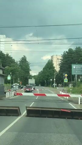 The video captures a train passing through a railway crossing in Kuzmolovskiy, Russia. The scene is filmed from the perspective of a vehicle stopped at the crossing, waiting for the train to pass. The sky is overcast, suggesting a cloudy or potentially stormy day. The railway crossing is equipped with a barrier gate, which is down, preventing road traffic.

The train is a modern, red and silver passenger train, identified by the Cyrillic text "Ласточка" (Lastochka, meaning Swallow) on its side. The word "РЖД" (RZD, Russian Railways) is also visible, along with a train number. Passengers can be seen through the windows of the train, suggesting it is in operation. The train tracks run across the road, and the crossing area includes a raised section with wooden planks.

In the background, there are trees, buildings (including apartment blocks), and overhead power lines for the train. A road sign with Russian text, "ОХОТНИЧЬЯ" (Okhotnichya, meaning Hunter's), is visible on a green billboard to the right, indicating a possible street or area name. A pedestrian crossing sign is also present. The overall impression is a typical urban or suburban railway crossing encounter.