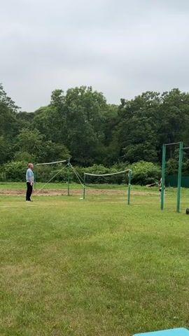 The video shows two people playing badminton on a grassy field. In the foreground, a man in a light blue shirt and dark pants stands on the left side of the court, holding a badminton racket. On the right side of the court, a younger person in a light gray top and dark pants is also holding a racket, ready to hit the shuttlecock. The net is set up in the middle of the field, with green poles supporting it. The background is filled with lush green trees, suggesting a natural or park-like setting. The sky appears overcast, indicating a cloudy day, possibly with a chance of rain. The time of day seems to be daytime, given the ambient light. The overall atmosphere appears casual and recreational, with the two individuals engaged in a friendly game of badminton. The location is identified as Fokino, Russia. There is no visible text in the video.