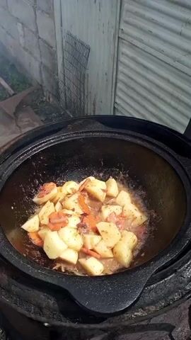 The video shows a close-up view of food being cooked in a large, black, cast-iron pot outdoors. The pot is placed over a fire, with embers visible at the bottom. Inside the pot, chunks of potatoes and sliced carrots are being stir-fried in oil. There are also pieces of meat and spices mixed in with the vegetables. Steam rises from the pot, indicating that the food is hot and actively cooking. The background is slightly blurred, showing a white wall and a corrugated metal door or fence. The overall scene suggests a rustic outdoor cooking setting, possibly a barbecue or a traditional meal preparation.