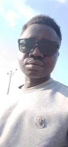 A close-up shot of a man with dark skin, a mustache, and wearing dark sunglasses. He has short, dark, textured hair. He is wearing a gray long-sleeved shirt with a small silver-colored emblem on the chest. The background shows a bright blue sky and the tops of buildings and power lines. The camera appears to be moving slightly.