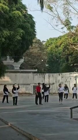 A group of approximately eight individuals, some wearing hijabs, are engaged in an outdoor activity on a paved surface. They are spread out across the frame, with several individuals holding long, thin objects, possibly sticks or poles. One individual in the center is wearing a red shirt and black pants and appears to be leading or instructing the group. The activity seems to involve coordinated movements, as suggested by the arm gestures of several participants. The environment includes trees and a building with a white wall in the background. The lighting indicates it is daytime, possibly early morning or late afternoon.