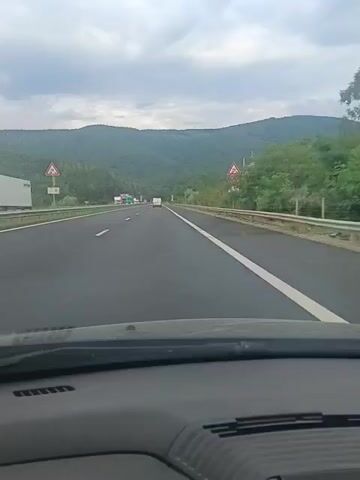 The video shows a view from the inside of a car as it travels on a highway. The highway is paved and has multiple lanes. Traffic signs are visible on the side of the road. The car is driving through a hilly, wooded area. The sky is cloudy. The dashboard of the car is visible in the foreground.