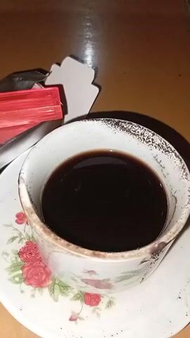 A close-up video shows a cup of black coffee. The cup has a floral design with red roses. The rim of the cup appears to have some dark residue. Next to the cup, partially visible, is a small red object which resembles a pack of candies or gum. The background is a warm, brown surface.