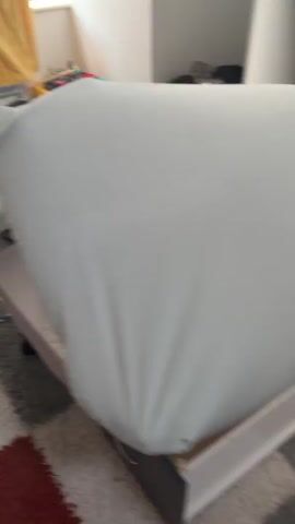 The video opens with a close-up of a white sheet covering a bed. A white stool with a chrome base is visible to the left. The camera pans to the right, revealing a black oscillating fan. The perspective then shifts to show a person lying on a bed, holding a tablet and a bottle of water. The environment appears to be a bedroom with carpeting and a woven laundry basket in the background.