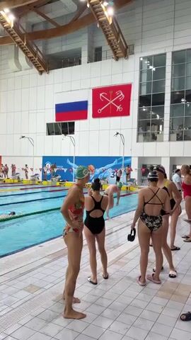 The video shows an indoor swimming pool area with multiple swimmers preparing to compete or having just competed. Several individuals are standing by the pool, some in swimsuits and swim caps. One person in the foreground on the left is adjusting their earbuds while looking towards the pool. In the background, several lanes of the swimming pool are visible with swimmers in them, and a large digital scoreboard is displayed above the pool. There are flags hanging on the wall, including the Russian flag and another flag with a red cross and anchor symbol. The environment appears to be a sports facility, likely a competition venue.