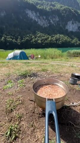 A close-up shot shows a pot of food cooking on a portable camping stove. The pot is made of metal and has a blue handle. The food inside appears to be a mixture of grains and possibly meat or vegetables. In the background, a blue tent is set up near a body of water, likely a lake. Tall green reeds grow along the shore of the water. Beyond the lake, a lush green forest covers the side of a steep mountain. The sky is clear and blue, indicating a sunny day. The overall scene depicts a camping or outdoor cooking scenario in a scenic natural environment.