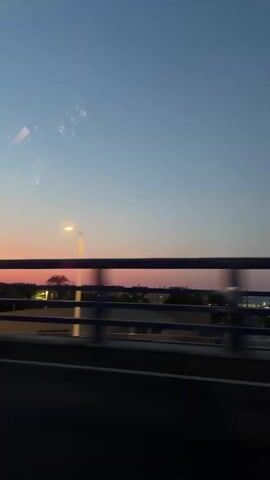 The video shows a view from a moving vehicle at dusk. The sky transitions from a clear blue at the top to hues of pink and orange near the horizon. A streetlamp is visible in the mid-ground, illuminated. In the background, there is a low-rise urban landscape with some trees, including palm trees. A highway railing or barrier is in the foreground, and a dark road surface is visible. The overall impression is one of a sunset viewed from a bridge or elevated road.