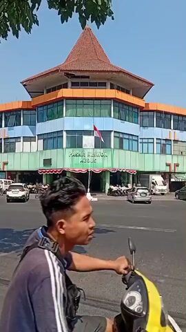 The video shows a street scene in front of a multi-story building with a distinctive orange and red tiled roof. The building has several floors with colorful facades in shades of green, blue, and orange, and many windows. A flag of Indonesia is displayed prominently on a flagpole in front of the building. The text "PASAR KLIWON KUDUS" is visible on the lower part of the building. The street in front is paved and appears to be an intersection. There are several vehicles, including cars and motorcycles, on the street. Some people are visible near the entrance of the building. A black pickup truck with a blue tarp covering its bed is driving across the street. The sky is clear and blue, indicating a sunny day. The overall environment appears to be a bustling urban or semi-urban area.