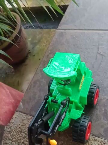 A green toy tractor with red wheels is lying on its side on a tiled surface. The tractor appears to be a front-end loader with a movable arm. The background shows some foliage and gravel. The camera is positioned low, at the level of the toy.