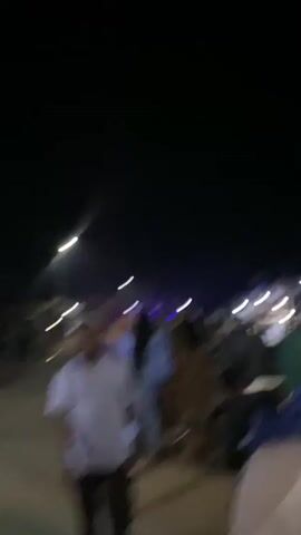 The video shows a nighttime scene on a paved street, illuminated by streetlights and possibly portable lighting. A crowd of people are walking or standing, some wearing light-colored clothing. A motorcycle with a rider is visible in the middle ground. In the foreground, the back of a person wearing a green garment is visible. There are buildings or structures lining the street. Blue light beams emanate from somewhere in the scene, casting a purplish hue. The overall impression is of a lively outdoor gathering or event at night.