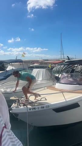 A man in a teal t-shirt and white shorts is on the deck of a white boat in a marina. He is bending over and appears to be adjusting something on the bow of the boat. The boat has a registration number "6º PM-1-86-25" visible. Other boats are docked nearby, some covered with white tarps. The sky is blue with scattered white clouds. The sun is shining.