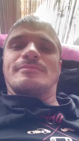 The video is a selfie-style recording of a man, likely in his late 20s to early 40s, with short, dark hair and a stubble beard. He is lying down, possibly on a couch or bed, and the camera is positioned above him, looking down. The background appears to be an indoor setting with what looks like blinds or curtains behind him, suggesting it might be daytime.

The man is wearing a dark t-shirt with a graphic design. Some of the text on the t-shirt is partially visible and appears to be in pink or red lettering. Phrases like "DAD" and "FOREVER" can be discerned, along with an image that looks like a game controller or console.

Throughout the short video clip, the man's facial expressions change. He starts with a neutral or slightly pensive look, then smiles subtly, and finally, he raises his right hand to flash a peace sign, with three fingers extended. His demeanor seems relaxed and casual. The overall tone of the video is informal, as if he is recording a short message or sharing a quick moment with someone. There are no specific cues about the time of day, weather, or precise location beyond the general indoor setting.