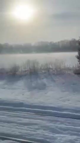 The video shows a sunny winter landscape. The sun is bright in the sky, shining down on a snowy field. In the distance, there is a light fog over what appears to be a body of water, with some tree lines visible through the mist. The camera appears to be moving alongside the snow-covered ground.