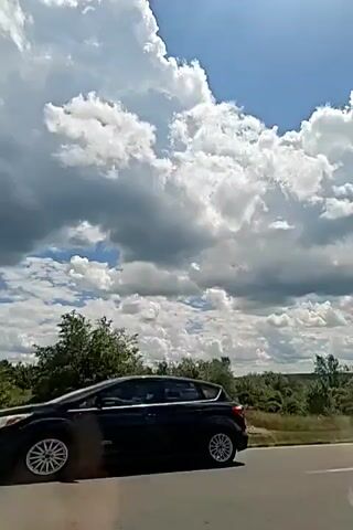 A video recording captures a view from a vehicle traveling along a road. The sky is predominantly blue with large, fluffy white clouds, some of which are shaded gray, suggesting potential for rain. Below the sky, a dense line of green trees and vegetation stretches across the horizon. The foreground shows a portion of the road, with a gray asphalt surface visible. The camera appears to be angled slightly upwards, focusing on the sky and the trees. There are no people or specific objects clearly identifiable within the frame. The overall impression is a scenic drive through a natural landscape on a cloudy but bright day.