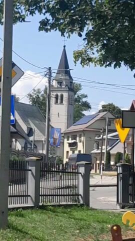 The video shows a daytime outdoor scene. A tall, narrow church tower with a pointed steeple dominates the background, with trees and power lines in front of it. A building with solar panels is visible to the right of the tower. In the foreground, there is a metal fence and some green grass. Several traffic signs are also visible, including a yellow diamond-shaped sign with a black border and a white arrow pointing to the right, and a white rectangular sign with a black border and a black arrow pointing left. The sky is blue and clear. There are no people or animals visible in the frame.
