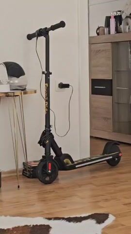 The video shows an indoor scene, starting with a black and gold electric scooter leaning against a white wall next to a small gold table with a white helmet on it. The scooter is plugged into an electrical outlet. The camera pans to the right, revealing a light wood and black cabinet with a TV mounted above it. The TV screen shows a video game with cartoon figures and a smaller image of a person wearing headphones and a microphone. The floor is covered with light brown wood flooring with a brown and white rug. The overall environment suggests a modern living room setting.