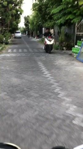 A person on a dark-colored scooter rides down a paved road lined with trees. The person is wearing a black shirt and shorts. The scooter has a white and red body and is moving forward, with the headlight illuminated. In the background, there are houses, more trees, and a white car parked on the left side of the road. A pedestrian crossing is visible on the road ahead. The timecode indicates the beginning of the video.