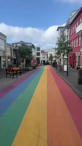 Here's a description of the video:

The video shows a pedestrian street that has been painted with a large rainbow running its length.  The colors of the rainbow are distinct and bright, taking up most of the width of the street. 

On either side of the rainbow pathway are sidewalks lined with buildings. Some buildings are painted in vibrant colors like red, while others are white or more neutral. There are trees planted along the sidewalks, and some outdoor seating areas with tables and chairs. A few people are walking in the distance along the rainbow-painted street.

The sky above is blue with some fluffy white clouds. The overall impression is of a bright, colorful, and welcoming urban environment.