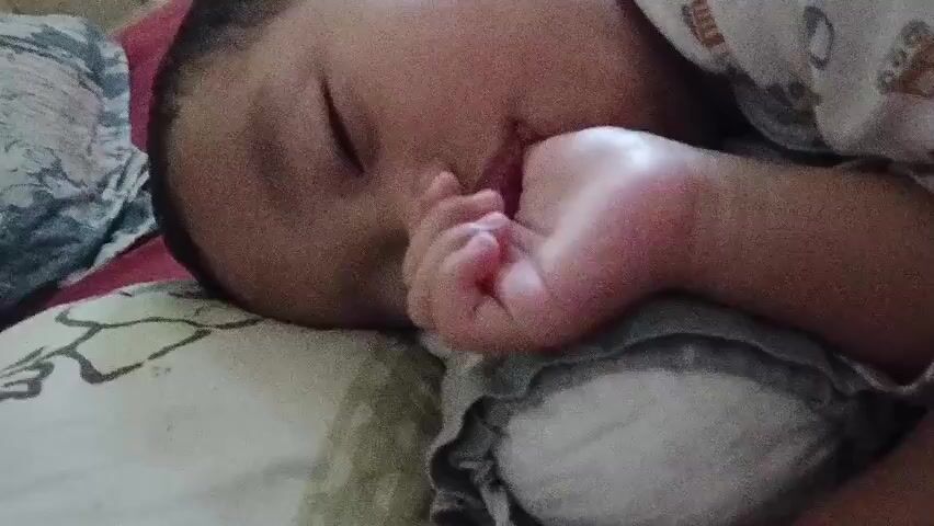 A close-up video shows a baby sleeping on its side with its mouth open and its hand near its mouth. The baby is wearing a white shirt with a cartoon pattern. The baby is on a pillow with a green and white pattern. The background is blurry, but appears to be a bed.