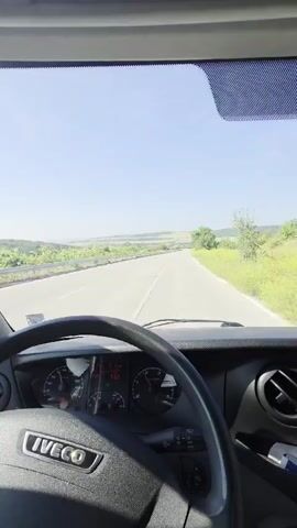 The video provides a first-person view from inside a vehicle, likely an Iveco van or truck, as indicated by the "IVECO" logo prominent on the steering wheel. The vehicle is traveling on a paved two-lane road that stretches into the distance under a bright, clear blue sky. Green vegetation, including trees and fields, lines both sides of the road. In the background, rolling hills covered in greenery are visible. The sun is shining brightly, suggesting daytime. The dashboard and various gauges are partially visible below the steering wheel. The scene depicts a calm drive through a scenic, rural landscape in Gamzovo, Bulgaria.