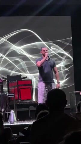 A man is on a stage holding a microphone and speaking. He is wearing a dark short-sleeved polo shirt and light-colored patterned pants. Behind him is a large screen displaying white wavy lines. To his left are some red boxes stacked on a stand. The audience is visible in the foreground, mostly in silhouette.