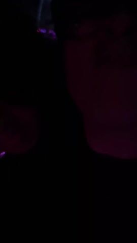 The video shows a person in glasses and a hat at a club with music and people dancing. The scene is very dark, with colorful strobe lights illuminating the crowd. People are holding drinks and enjoying the music.