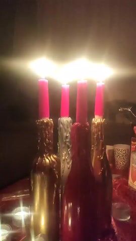 Four wine bottles are used as candle holders, each with a pink candle burning atop. The bottles are decorated with dripping wax, painted gold, silver, and red. They sit on a red tablecloth, surrounded by small tea lights. The lighting is dim and warm, and there is music playing in the background.