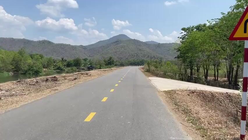 The camera moves slowly along a paved road with yellow dashes. The road is flanked by a body of water on the left and sparse vegetation on the right. In the distance, a mountain range is visible under a sky with scattered clouds. There appears to be some construction or disturbance of the land near the water's edge.