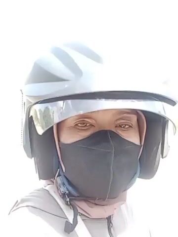 A person wearing a gray helmet, a black face mask, and a light pink headscarf is looking at the camera. The person is wearing a gray hoodie. The background is blurred, but appears to be outdoors with some trees visible. The person is stationary.