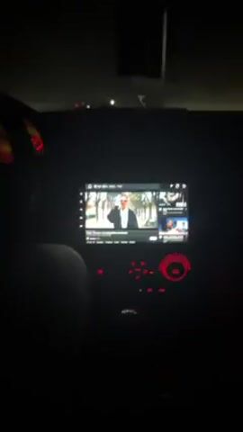 From the interior of a vehicle at night, the video shows a person driving, watching a music video on the car's entertainment system. The video is playing on YouTube, showing a man standing next to a luxury car in front of a photo of the Eiffel Tower. The car is driving on a dark road. The car's dashboard lights are red.