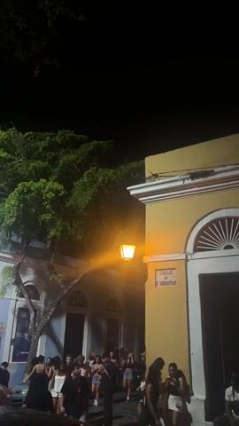 The video captures a nighttime street scene in what appears to be a historic urban area. The foreground is dominated by a yellow building with a white arched doorway. A street sign reading "CALLE 10 SEBASTIAN" is visible. Several people are gathered on the street, some appearing to be socializing outside buildings. The atmosphere seems lively, possibly indicating a nightlife or event. The background is dark, suggesting the presence of trees and other structures. The lighting is primarily from a street lamp and decorative string lights, creating a warm and festive ambiance.