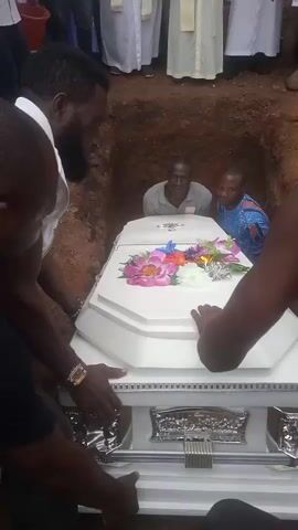 A group of people are gathered around an open grave. Two men are inside the grave, leaning over a white coffin adorned with flowers. One of the men in the grave appears to be crying or overcome with emotion. Other individuals are standing at the edge of the grave, some appearing to be wearing white robes, suggesting a funeral or religious ceremony. The ground around the grave is reddish-brown soil. The coffin is being lowered into the grave, with several people on the outside of the grave holding onto it. The overall atmosphere suggests a somber event.