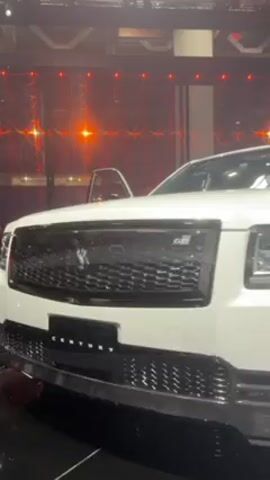 The video shows the front and side view of a white Toyota Century with black rims, a black grille, and a black front bumper. The car is sitting in front of a red and white patterned background. The camera slowly pans from the front of the car toward the front tire.