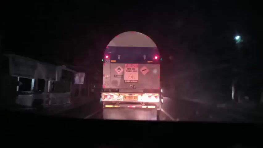 The video shows the back of a tanker truck on a road at night. The truck has various warning signs on its back, including a “Blind Spot” warning. The road has a yellow line down the center. The surrounding area appears to be a residential or commercial area with buildings visible on the sides of the road. The video is taken from the perspective of someone driving behind the truck. The weather seems clear.