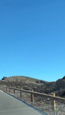The video shows a roadside view of a barren, rocky landscape under a clear blue sky. A paved road with a white line marking the edge curves to the left. A wooden fence with vertical posts and horizontal rails runs alongside the road, separating it from the rocky terrain. The landscape is characterized by dark, irregular volcanic rocks and sparse vegetation. The camera appears to be moving, as the perspective shifts slightly and the fence posts become more distinct. The overall impression is of a desolate, natural environment, possibly a volcanic park or a remote area with volcanic activity.