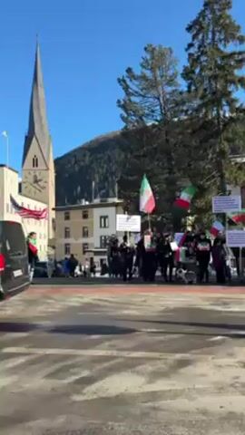 The video shows a group of people protesting in a town square on a sunny day. Some people are holding Iranian flags and signs, and an American flag is also visible. The protesters appear to be chanting.  There is a church with a tall spire in the background, along with other buildings. The ground has some patches of ice or snow. Cars are driving by on the street in front of the protesters.