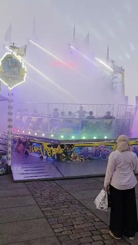 The video shows a carnival ride in operation. The ride features multiple cars that spin and lift riders into the air. Lights and fog are used to create a dramatic effect. A woman stands in the foreground, watching the ride. The ground is paved with bricks, and there is a metal ramp leading up to the ride. The sky is overcast, suggesting it may be evening or a cloudy day.