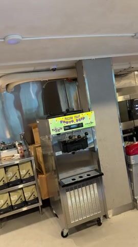 The video shows a close-up of a commercial soft-serve ice cream machine. The machine is stainless steel with a black dispenser head. A colorful sign is attached to the front of the machine that reads "PASSION FRUIT FROYO DRIP BY COCONUT CULT." Below the sign, there is a digital display and several handles for dispensing the froyo. The machine appears to be in a commercial kitchen or food service environment, as indicated by the industrial shelving and equipment visible in the background. The lighting is bright and even, suggesting an indoor setting.