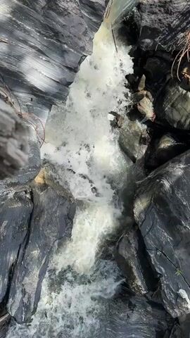 This is a video recording looking down at a fast-flowing river cascading over rocks. The water is white with foam, indicating a strong current. The river is surrounded by large, dark gray rocks, some with striations suggesting metamorphic or sedimentary origins. The lighting suggests it is daytime, with sunlight casting shadows on the rocks. There are some sparse green plants growing on the rocks in places. The scene is natural and wild. There is no visible text in the video. The overall impression is one of raw natural power and the beauty of a rugged landscape.