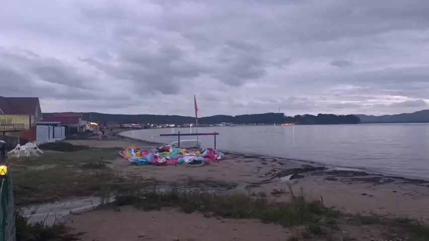 This is a static, eye-level video of a beach scene at dusk. In the foreground, there is dry grass and sand. In the middle ground, there is a pile of deflated inflatable toys, some of which are brightly colored. There is a small wooden structure with a red flag attached to it, and some white plastic chairs nearby. To the left, there are two small buildings with lights on. The beach curves into the distance, where there are more buildings and lights along the shoreline. The water is calm, and the sky is cloudy and overcast. The overall impression is one of a quiet, end-of-day beach setting.