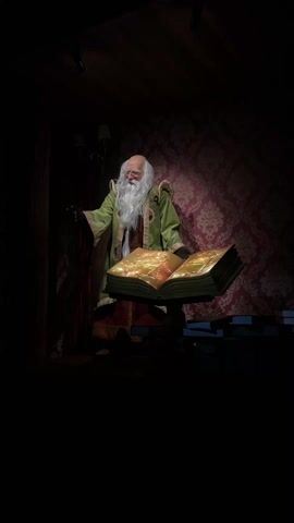 A close-up shot shows a figure resembling Santa Claus standing beside an open book. The figure has a long white beard and is dressed in a green robe trimmed with gold. The figure is partially illuminated by a spotlight, with a red patterned wall behind it. The open book on a stand glows with light, with an image on the pages. The room is dim, but a portion of a lamp is visible in the upper left corner. The overall aesthetic is festive, with a focus on fantasy and storytelling.