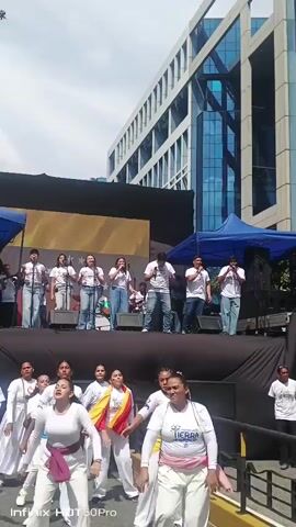 A group of singers performs on a stage with dancers in front of them, outside a modern building. The singers, dressed in white t-shirts and jeans, are lined up on the elevated stage, holding microphones. The dancers, wearing white outfits with pink and yellow sashes, are on the ground level, performing synchronized movements. The stage is set up outdoors with a dark backdrop and a large yellow banner featuring stars. A blue tent is visible behind the stage. The building in the background is tall and has a glass facade. The scene appears to be a public event or performance, possibly a festival or rally, with a crowd gathered to watch.