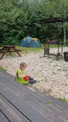 A young girl with blonde hair pulled into a ponytail, wearing a bright green t-shirt and grey leggings, sits on a pebble-covered ground. She is playing with a small orange bucket and a red shovel, seemingly engrossed in her activity. The setting appears to be an outdoor campsite.

In the background, a green tent is pitched, suggesting a camping trip. A wooden picnic table is visible to the left of the tent. To the right of the tent, there's a black metal structure with a canopy, possibly a barbecue grill station, with logs and grilling tools. Further to the right, a black trash bin stands. The ground is covered with small, light-colored pebbles.

The scene is surrounded by lush green trees, indicating a natural, wooded environment. The sky is overcast, and the lighting suggests it is daytime, possibly a cloudy afternoon. The overall atmosphere seems peaceful and relaxed, with the child enjoying a moment of solitary play. No text is visible in the media file. The location context provided is Verkhneye Dubrovo, Russia.